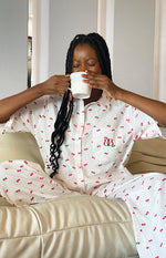 White cherry print pajama shirt with a red embroidered logo on the chest pocket and matching printed pants