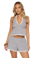 A studio shot from the neck to the waist of a woman wearing a halter-style top. This top features a small-scale plaid pattern in shades of purple and yellow. It has a plunging V-neckline and a straight hem both finished with a white eyelet lace scallop trim.