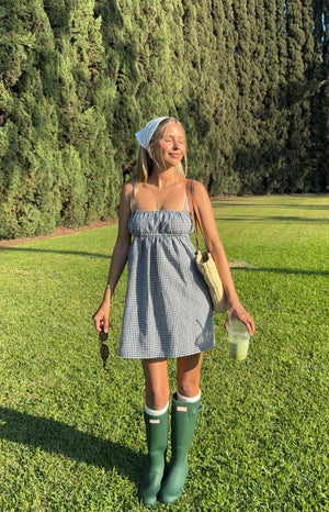 A woman in a black and white gingham mini dress with a ruched bust and spaghetti straps- accessorised with a white bandana- white socks- and dark green rain boots.