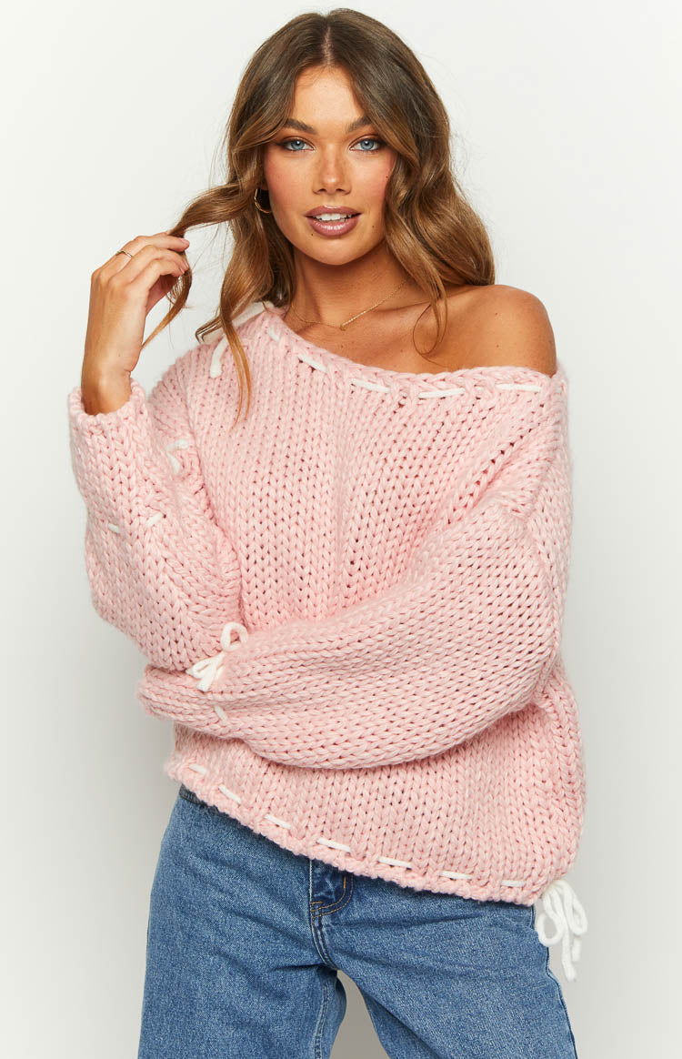 Baby pink chunky knit sweater with a wide off-shoulder neckline and white lacing details on the shoulders, sleeves, and side hem