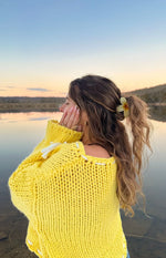 Rear view of a cropped yellow chunky knit sweater with white tie details on the sleeves and a curved hem
