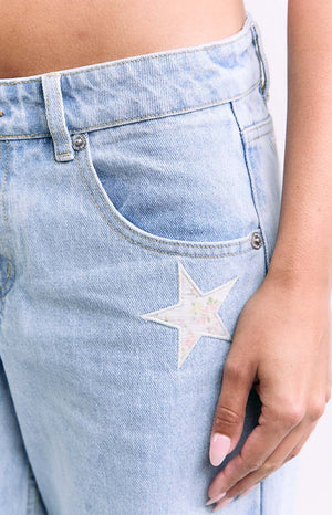 A close-up of the front pocket of light blue jeans featuring a small star applique with a subtle floral print pattern.