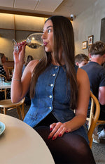 Side view of a mid wash denim button-down vest top featuring white buttons and a split hem