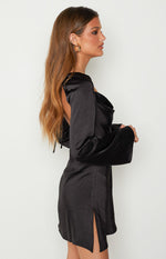 Side view of a black satin mini dress with a cowl neck, long bell sleeves, open back with tie detail, and a side slit