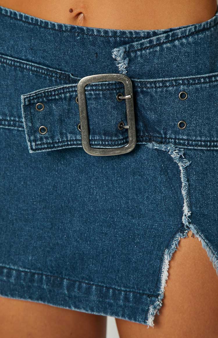 Side view of a blue denim mini skirt with a large silver buckle, eyelets, and a raw hem side split
