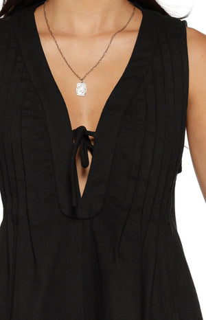 A lifestyle shot of our model wearing a short sleeveless black romper. This playsuit features a deep V-neckline with a keyhole opening that is secured at the top by a thin fabric tie. The bodice and flared shorts are accented with vertical pleated gathering.