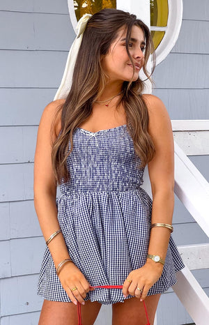 Model is wearing a navy and white gingham playsuit featuring a shirred bodice- thin shoulder straps- and a small white bow detail at the neckline. Her hair is accented with a cream bow.