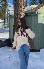 Angled front view of a cream chunky knit sweater with burgundy 'SKI' jacquard lettering and oversized long sleeves
