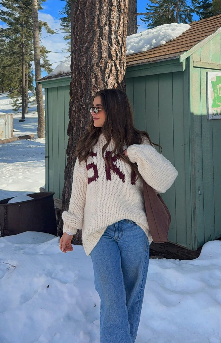 Angled front view of a cream chunky knit sweater with burgundy 'SKI' jacquard lettering and oversized long sleeves