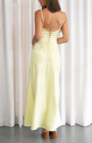 A rear view of a female model wearing a pale yellow satin maxi dress with a lace-up corset back detail.