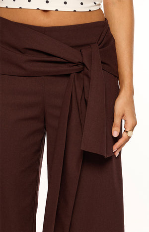 Our model wearing high-waisted flowy dark brown wide-leg trousers. This pants feature a large integrated fabric belt tied prominently into a knot at the front of the waist.