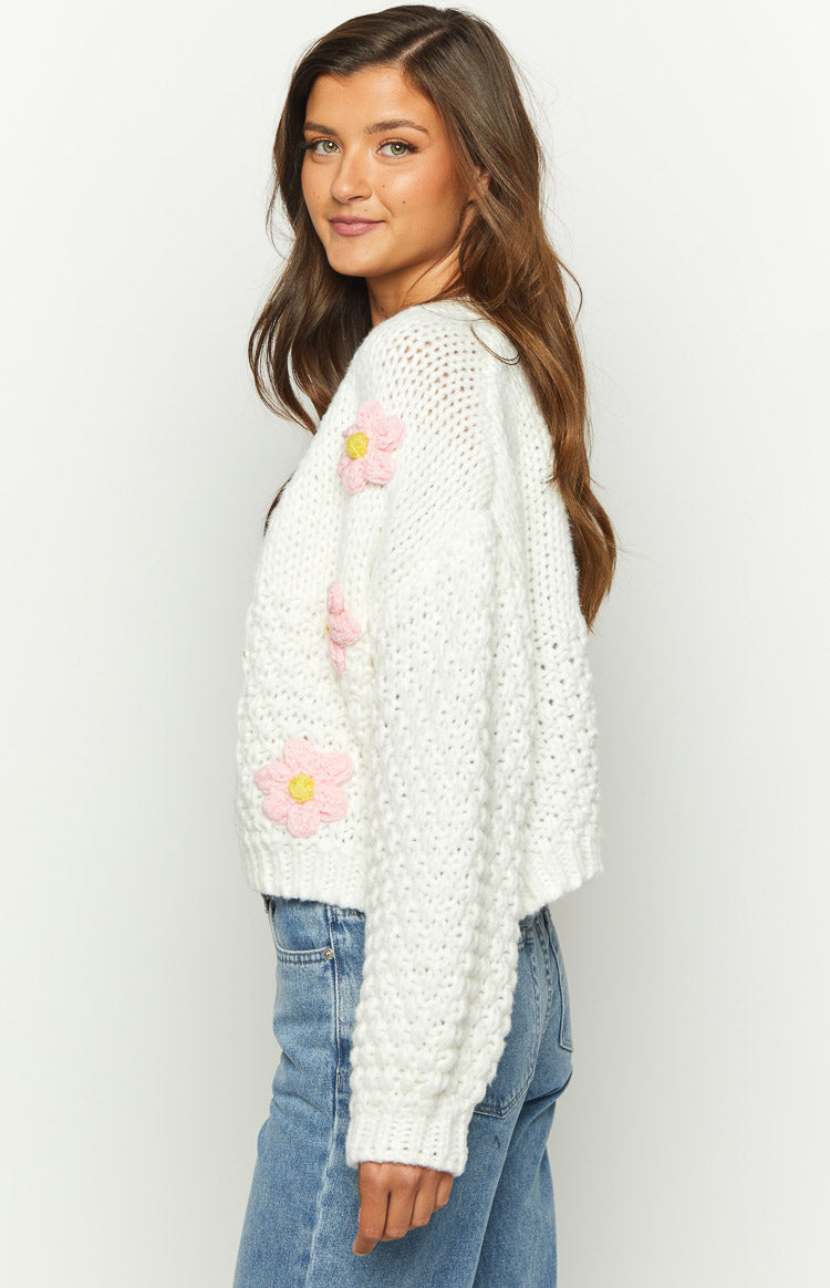 Side view of a white chunky knit cardigan with pink and yellow crocheted flower appliques on the sleeve
