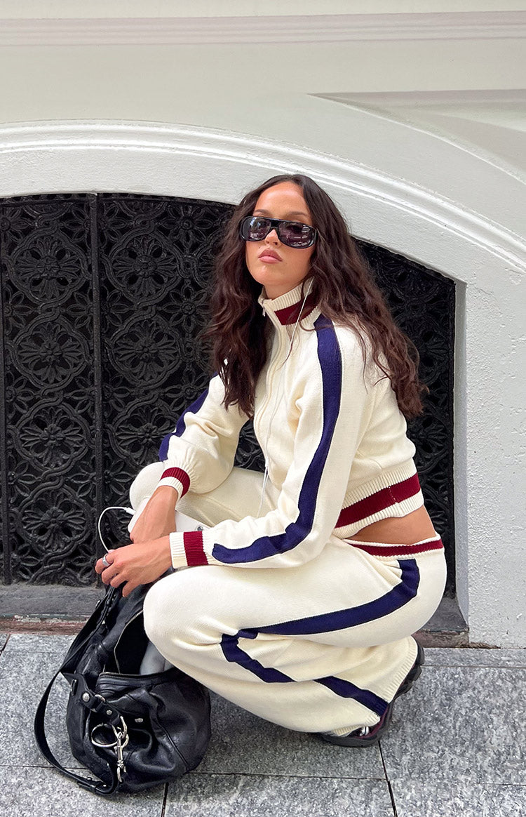Side view of cream knit jacquard track pants, showing the relaxed fit, dark blue side stripe, and ribbed ankle cuffs