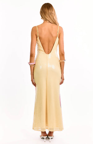 Back view of our model wearing yellow maxi dress with a deep V-shaped open back and thin spaghetti straps. This dress has a sheer shimmering sequin overlay with a subtle pink floral print on the side.