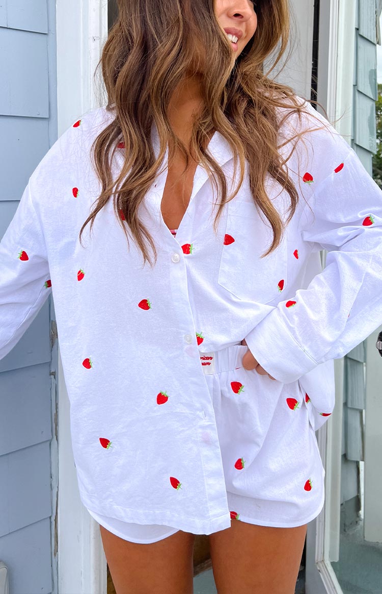 Model is wearing a white long-sleeve button-down shirt adorned with small red embroidered strawberries all over. The shirt is partially unbuttoned and paired with matching white shorts that also feature strawberry embroidery and a visible "Emmy x Beginning" label on the waistband.
