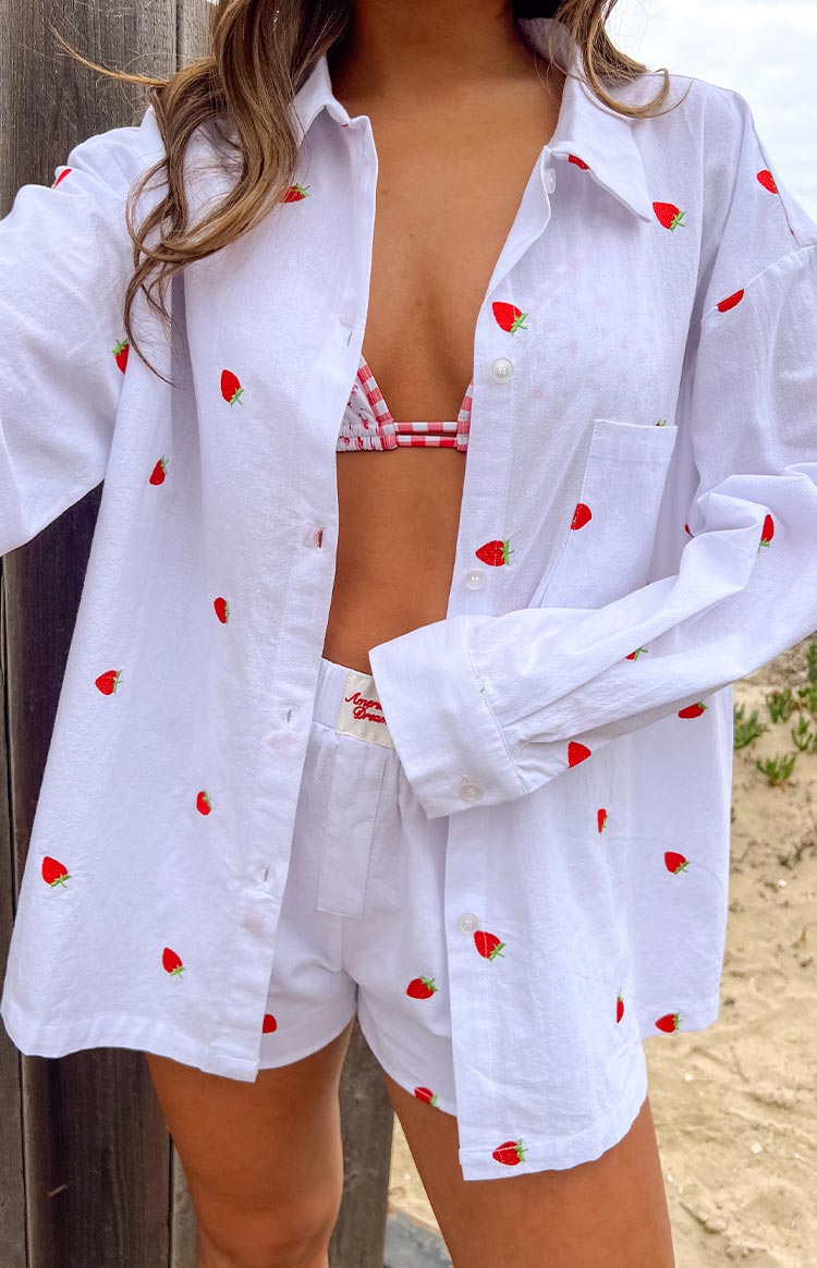 Girl is styled in a white oversized long-sleeve shirt with a scattered red strawberry embroidery pattern. The shirt is worn open- revealing a red and white gingham print bikini top- and is paired with matching white strawberry embroidered shorts.