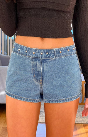 Close-up of mid-wash denim low-rise shorts- detailed with silver studs along the waistband- paired with a black long-sleeve top.