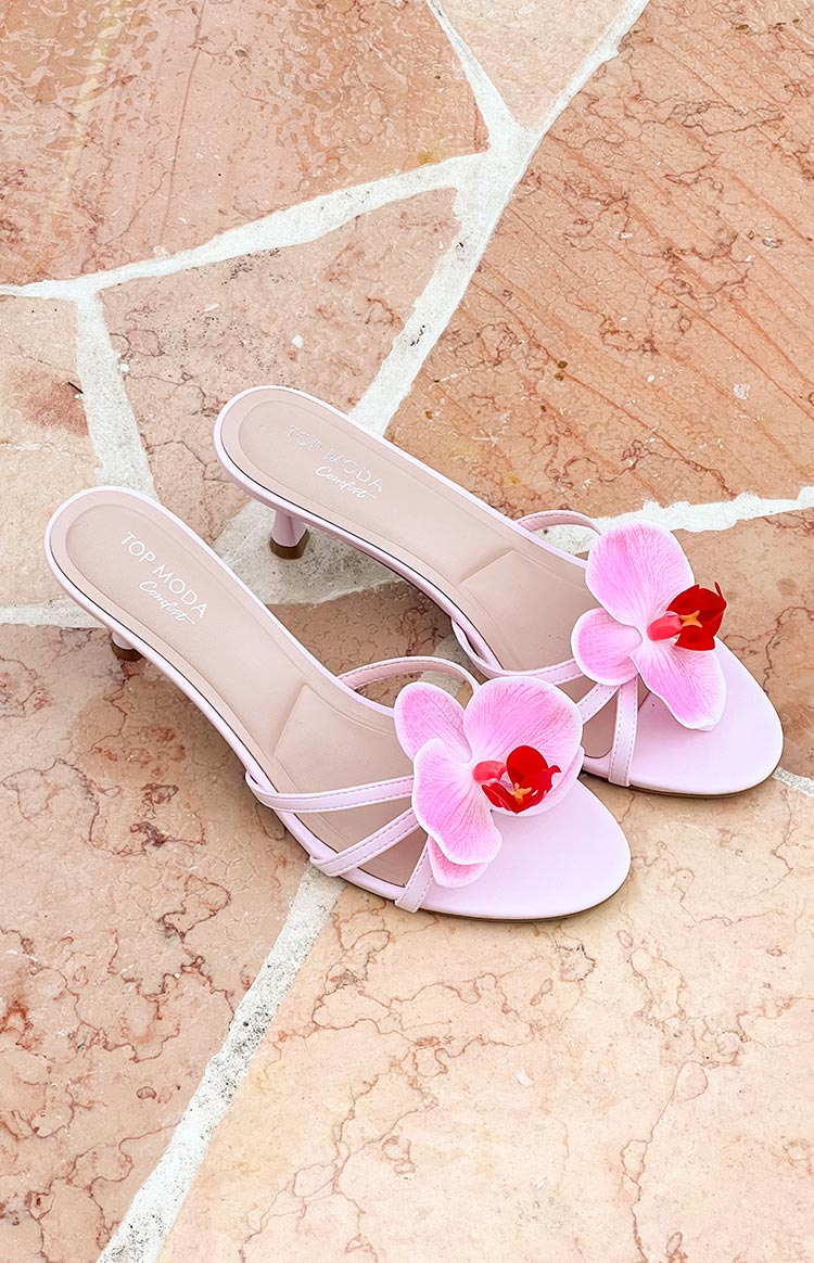 Two low-heeled sandals with orchid embellishments sit on a tiled surface. The sandals feature multiple thin straps.
