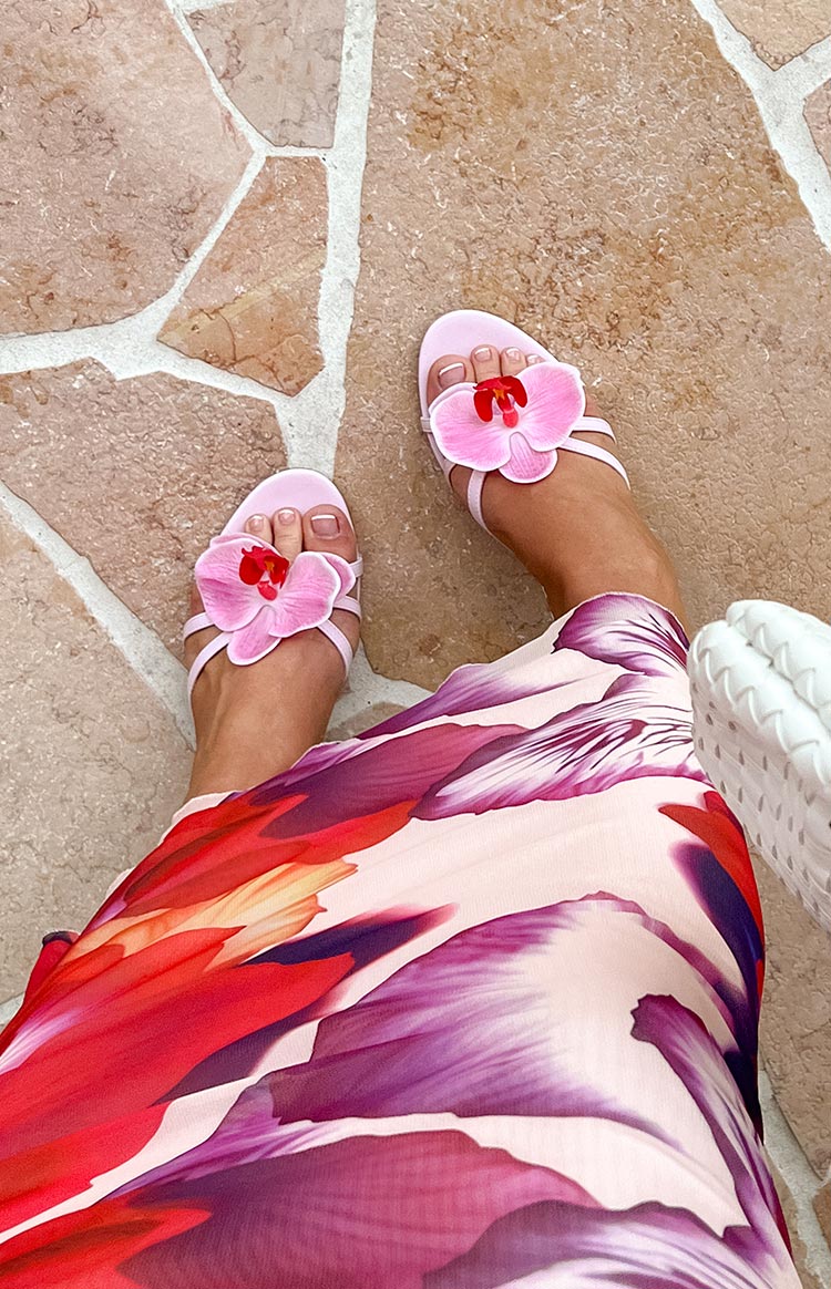 A person wearing low-heeled sandals with orchid decorations stands on a tiled surface. The person is also wearing a floral-patterned garment.