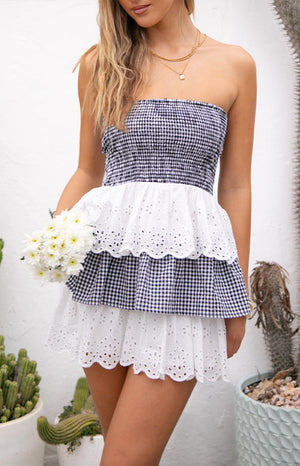 Model wearing a strapless mini dress with a navy and white gingham bodice and alternating white eyelet and gingham ruffled tiers on the skirt- holding white flowers.
