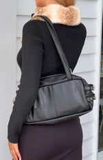 Side view of a black faux leather shoulder bag with two silver buckle straps on the side and a top zipper