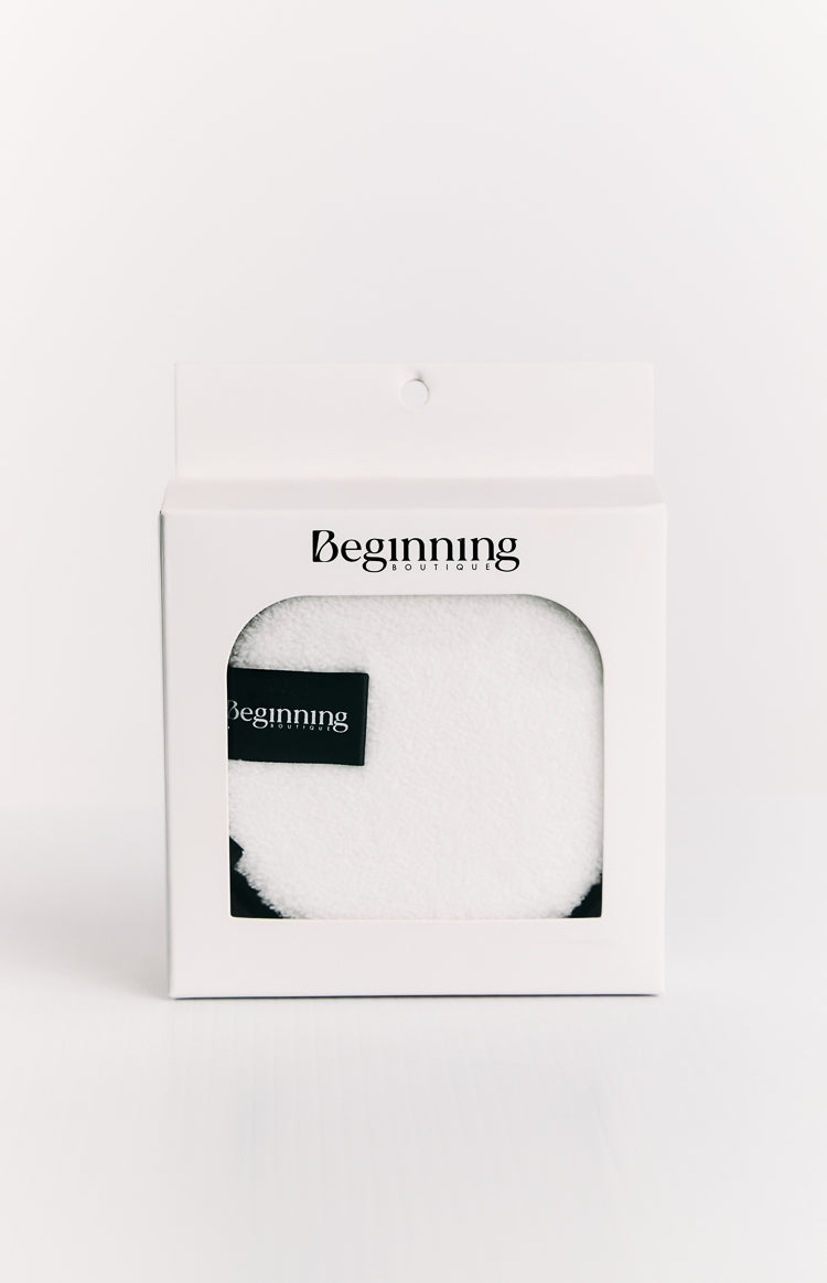 White fluffy reusable face wash pad with a black brand label, visible through the window of its white retail box
