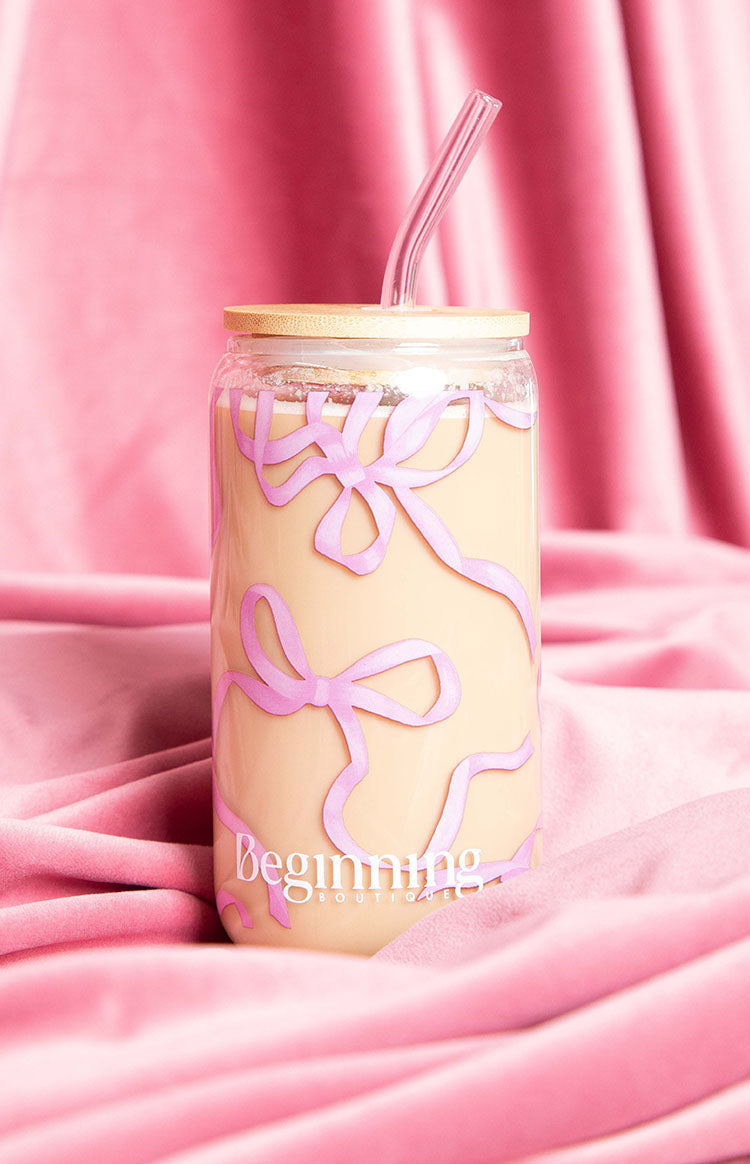 Glass tumbler with pink bow pattern, bamboo lid, and bent glass straw, filled with a creamy drink