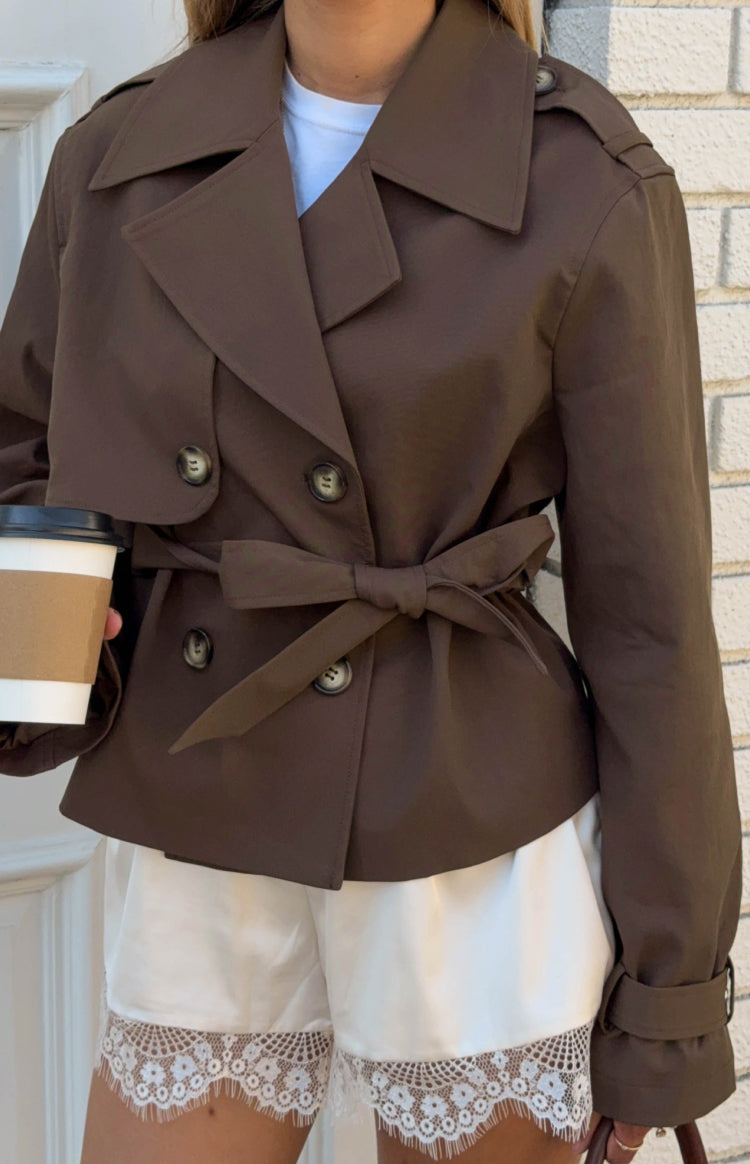 Close-up of a brown cropped trench coat with double-breasted buttons and a waist tie tied in a bow