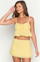 Brunette model wearing a yellow two piece set, flowy crop top and mini skirt with a gold necklace holding a drink.