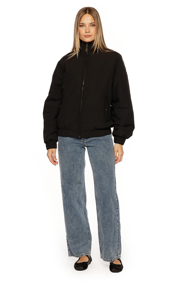 A close-up of our model wearing a black puffer bomber jacket with a relaxed slightly oversized fit. This jacket features a high stand collar and a prominent silver zipper down the front. It has an elasticized hem and cuffs with one hand tucked into a side pocket.