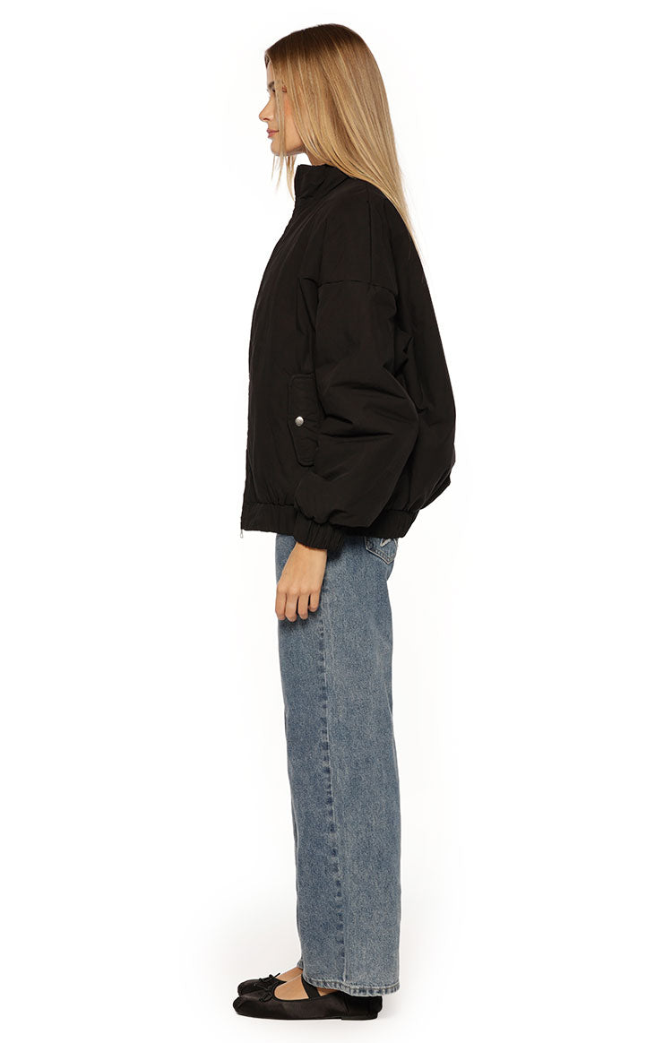 A close-up of our model wearing a black puffer bomber jacket with a relaxed slightly oversized fit. This jacket features a high stand collar and a prominent silver zipper down the front. It has an elasticized hem and cuffs with one hand tucked into a side pocket.