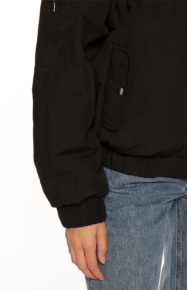 A close-up of our model wearing a black puffer bomber jacket with a relaxed slightly oversized fit. This jacket features a high stand collar and a prominent silver zipper down the front. It has an elasticized hem and cuffs with one hand tucked into a side pocket.