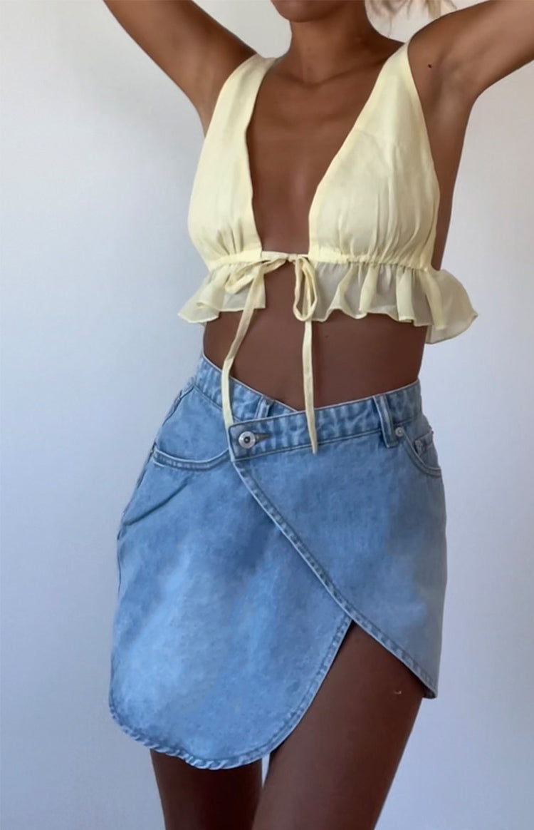 Light blue denim mini skirt with an asymmetrical wrap front, V-shaped waist, and single button closure