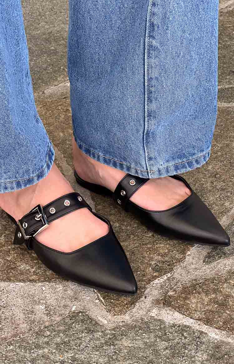 Black pointed-toe mules with a smooth finish, featuring a wide buckled strap across the foot with silver eyelets