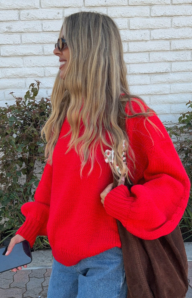 Side view of a red knit sweater with voluminous sleeves, long ribbed cuffs, and a small floral detail