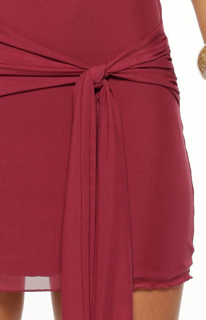 Our model wearing a fitted mini dress in a burgundy stretch fabric. This mini dress features an asymmetric off-the-shoulder neckline and a ruched side seam with a matching fabric tie knotted at the hip leaving a long sash hanging down the front of the short skirt.