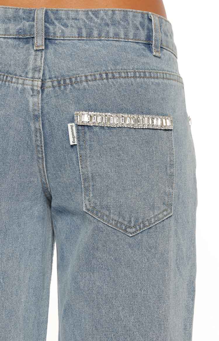 A close-up on the back of a pair of light-wash denim jeans specifically highlighting the right back patch pocket. The pocket is heavily embellished along the top edge with a row of clear rectangular baguette-cut rhinestones. The denim features a standard waistband with belt loops.