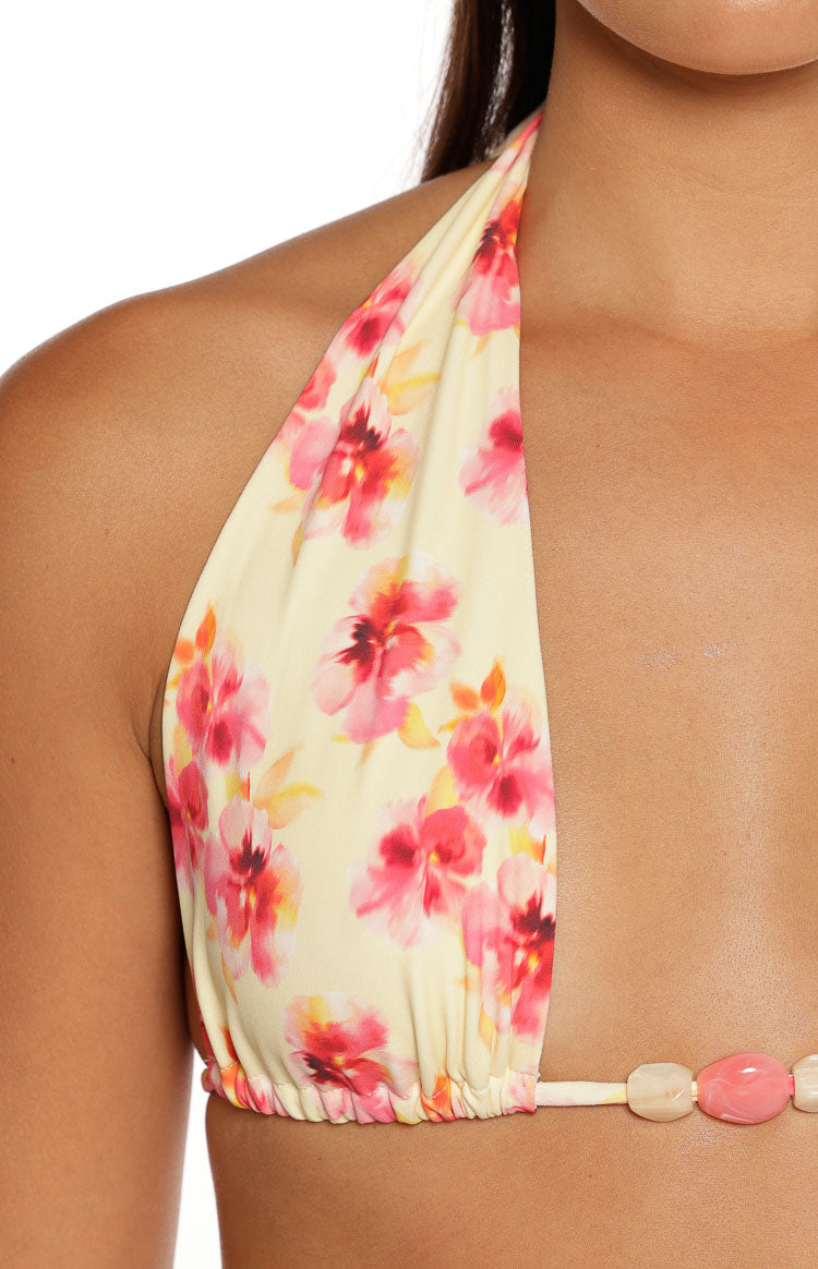 A tightly cropped close-up shows the right cup of a halter bikini top the fabric features a creamy yellow base printed with a vibrant pink and red watercolor-style floral pattern. The bottom edge of the cup is gathered and connects to a thin band that is decorated with a line of large decorative pink and white beads.